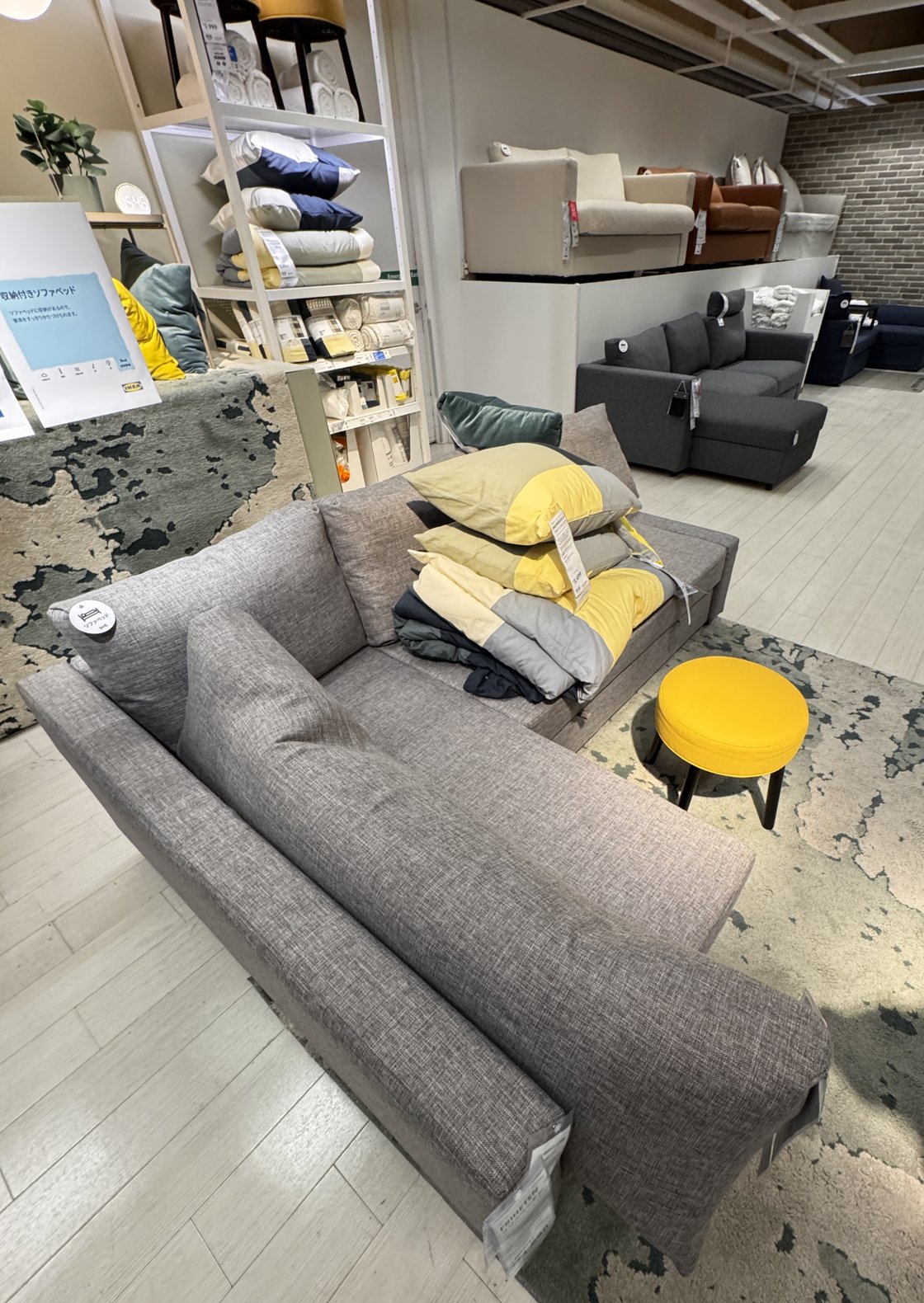 IKEAの売り場に置かれたグレーのL字のソファベッド。座面に黄色とグレーの寝具が積まれ、横に黄色のスツールがある