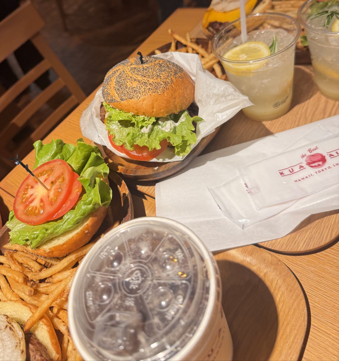 木のテーブルに並んだクアアイナのハンバーガー。ポピーシードのバンズのバーガー、トマトとレタスを別に盛ったバーガー、レモンの飲み物