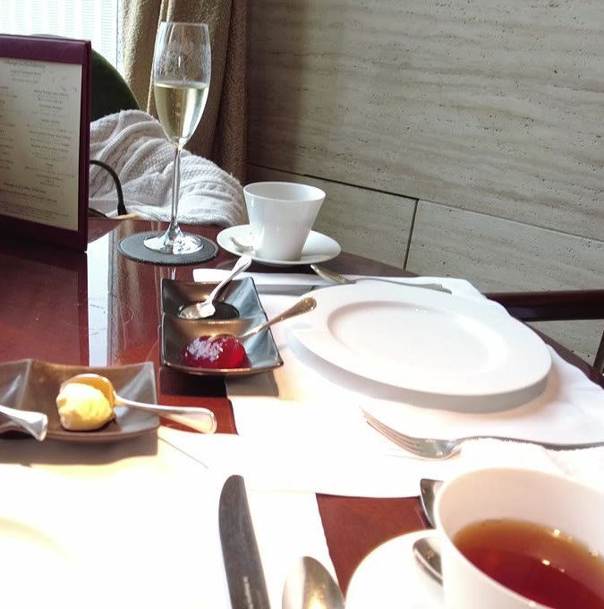 光沢のある木のテーブルに白い皿と紅茶のカップ。奥に石張りの壁と金色のカーテン