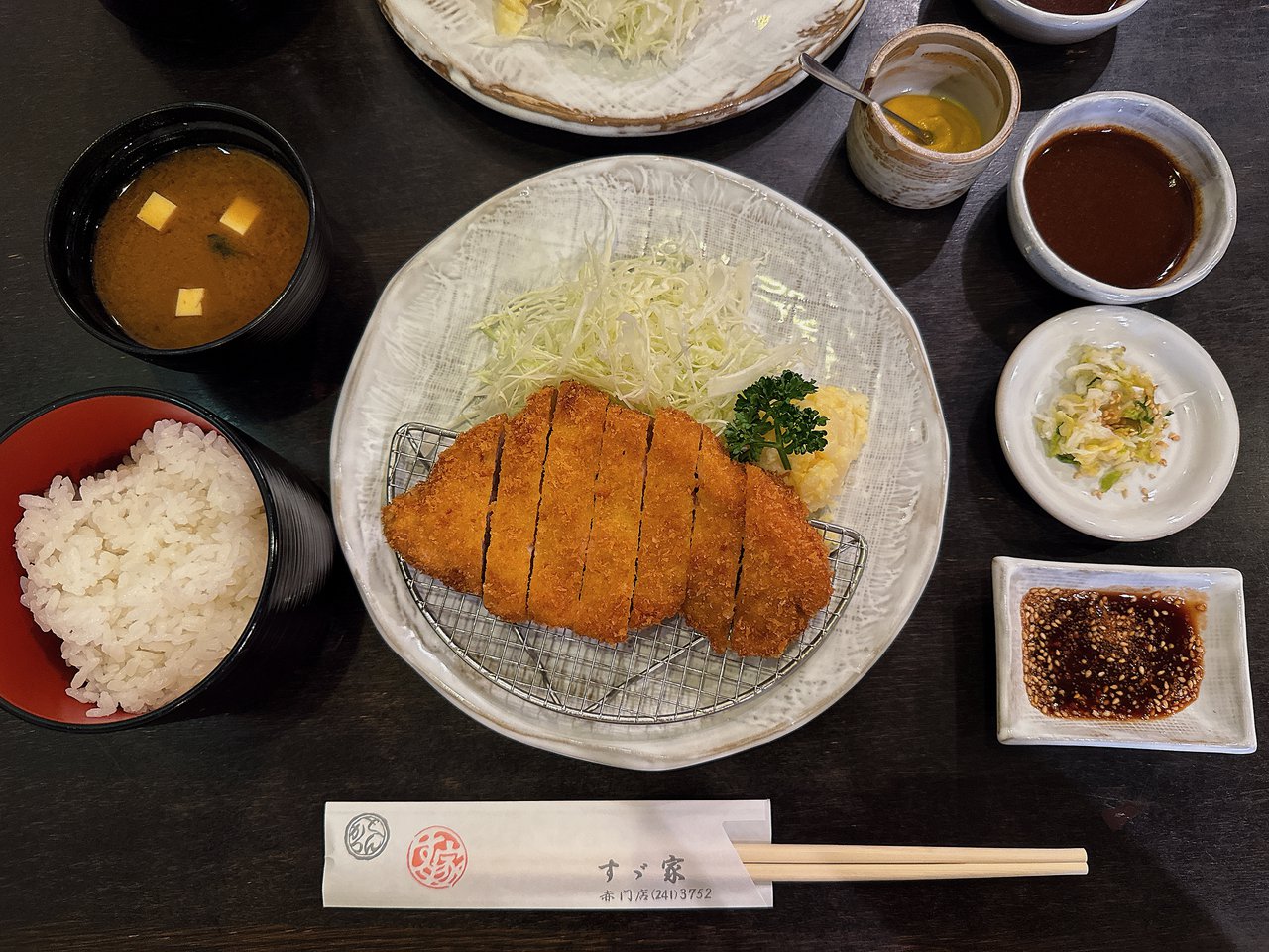 すゞ家 赤門店のとんかつ定食1人分。赤い椀のご飯・豆腐の味噌汁・からし・ソース・漬物・ごまの入ったたれ