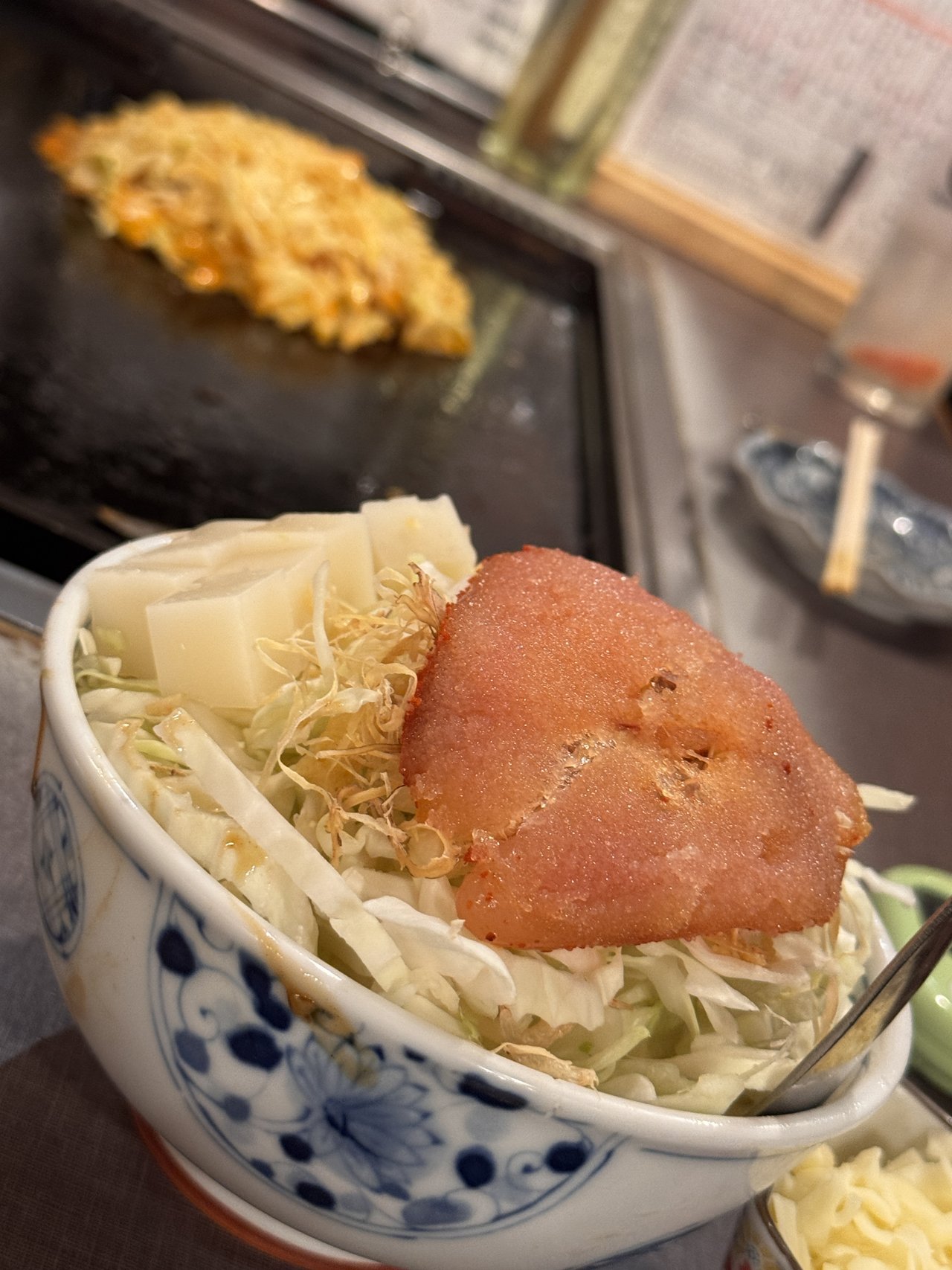 たまとやのもんじゃの具。染付の丼にキャベツと角切りの餅と明太子1腹がのり、手前に別皿のチーズ、奥の鉄板で先に焼いたもんじゃがのびている