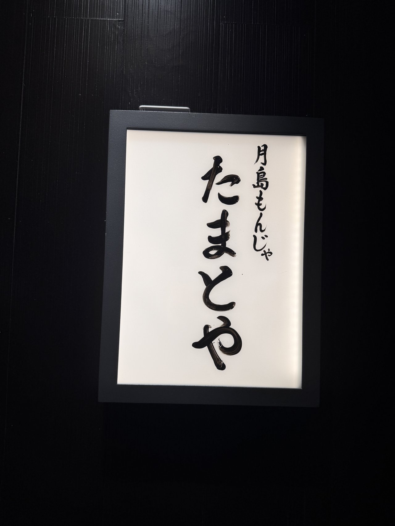 黒い壁にかかった白い看板。毛筆で月島もんじゃ たまとやと書かれている